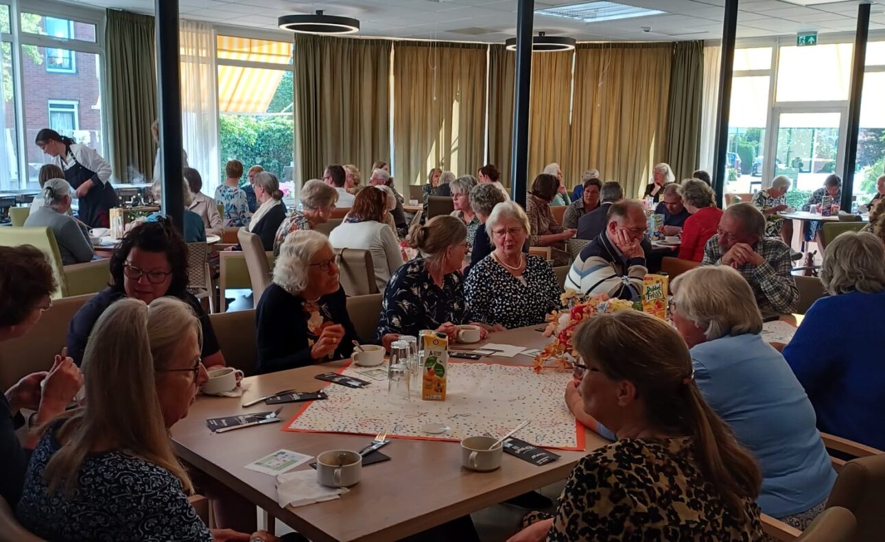 Groep mensen zit aan tafels in een lichte ruimte, genietend van een sociale bijeenkomst met koffie en gebak op tafel.