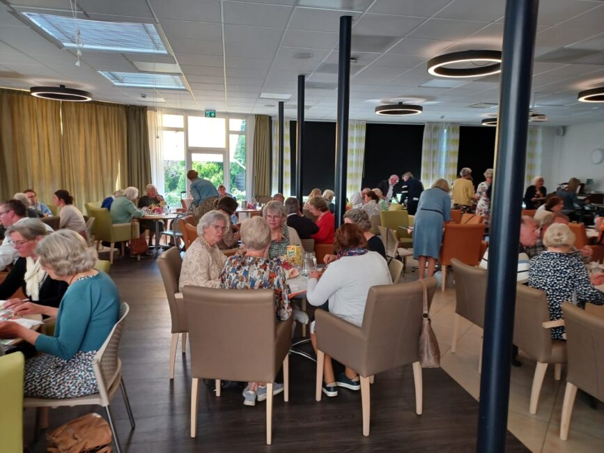 Een groep ouderen zit aan tafels in een gemeenschappelijke ruimte, druk in gesprek en genietend van een sociale bijeenkomst.