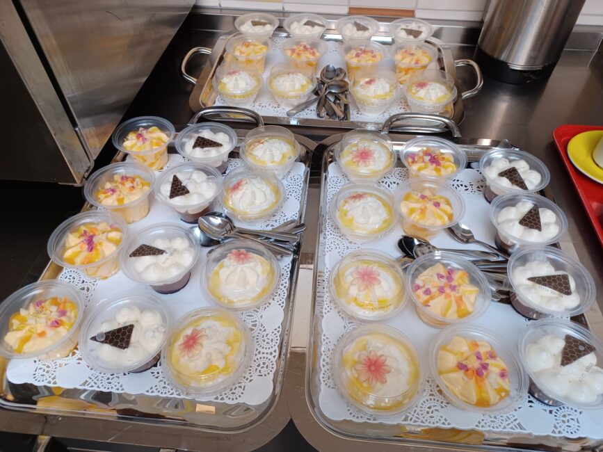 Vier dienbladen met desserts in plastic bakjes, versierd met slagroom, fruit en chocoladestukjes, op een metalen oppervlak.