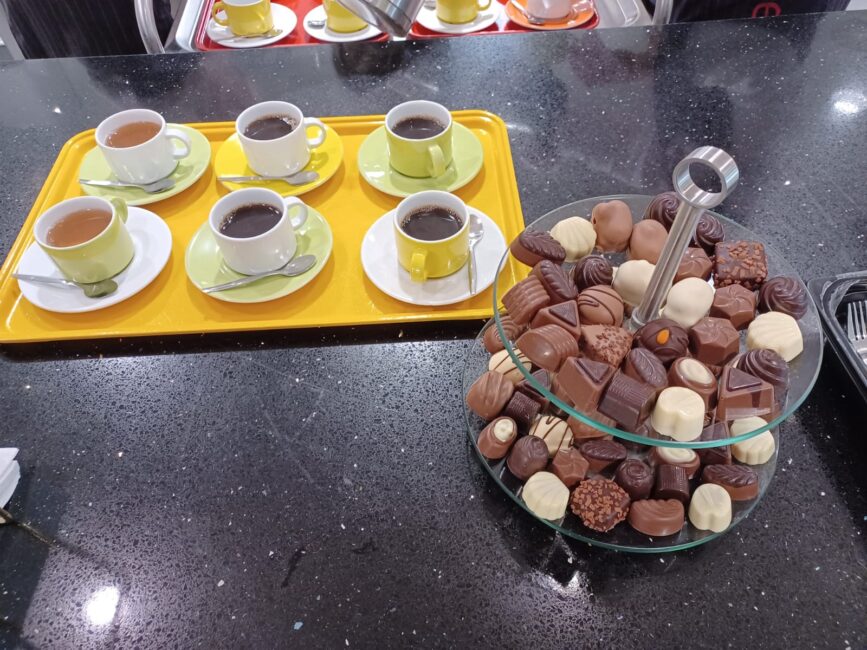 Een dienblad met zes kopjes koffie en thee naast een etagère vol met diverse soorten bonbons op een zwarte tafel.