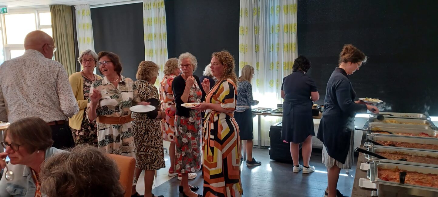 Groep mensen in een zaal, pratend en etend bij een buffet. Ze dragen kleurrijke kleding en de sfeer is ontspannen en gezellig.