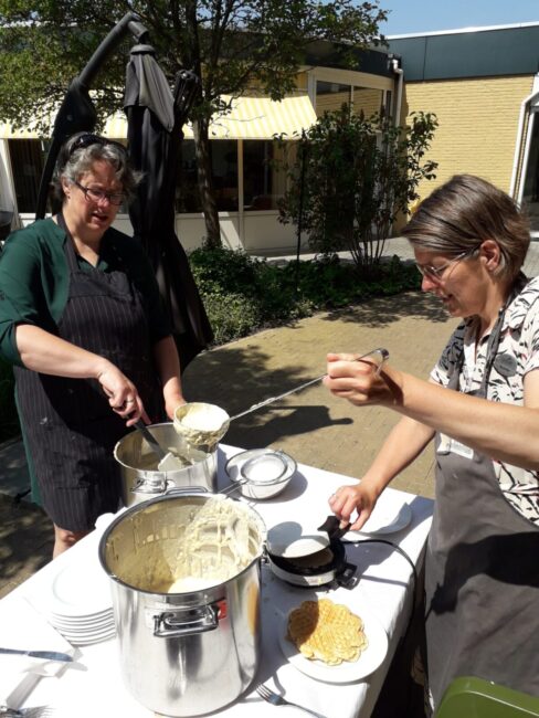 Twee vrouwen serveren eten buiten op een zonnige dag. Ze gebruiken een grote pan en scheppen het eten in kommen.
