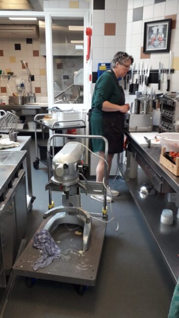 Een vrouw werkt in een professionele keuken, omringd door keukenapparatuur en gereedschap, met een grote mixer op de voorgrond.