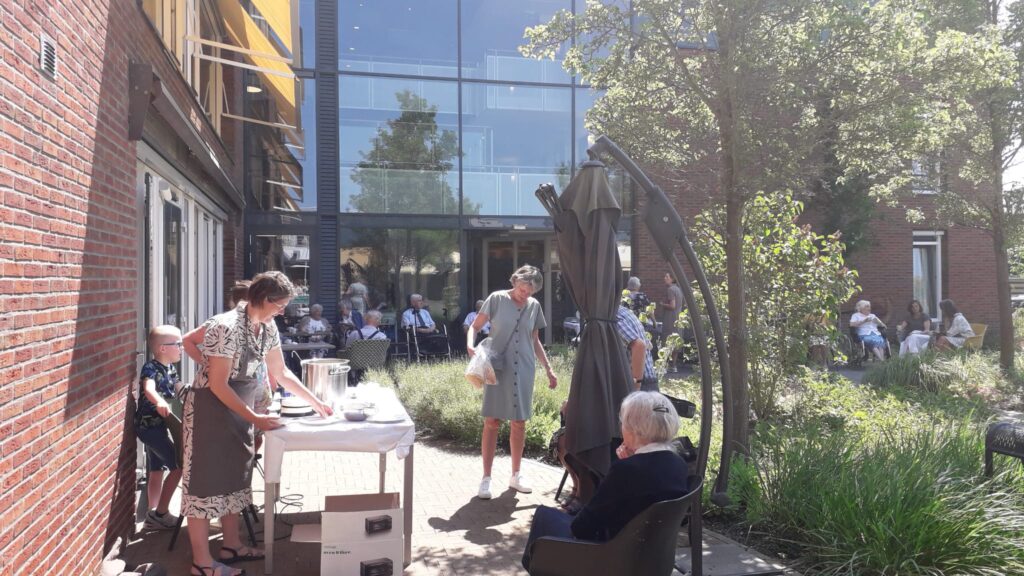 Mensen genieten van een zonnige dag buiten bij een zorginstelling, met tafels en stoelen in een groene omgeving.