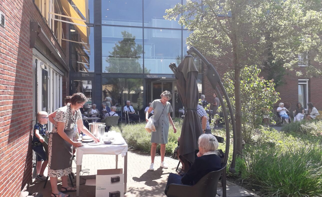 Mensen genieten van een zonnige dag buiten bij een zorginstelling, met tafels en stoelen in een groene omgeving.