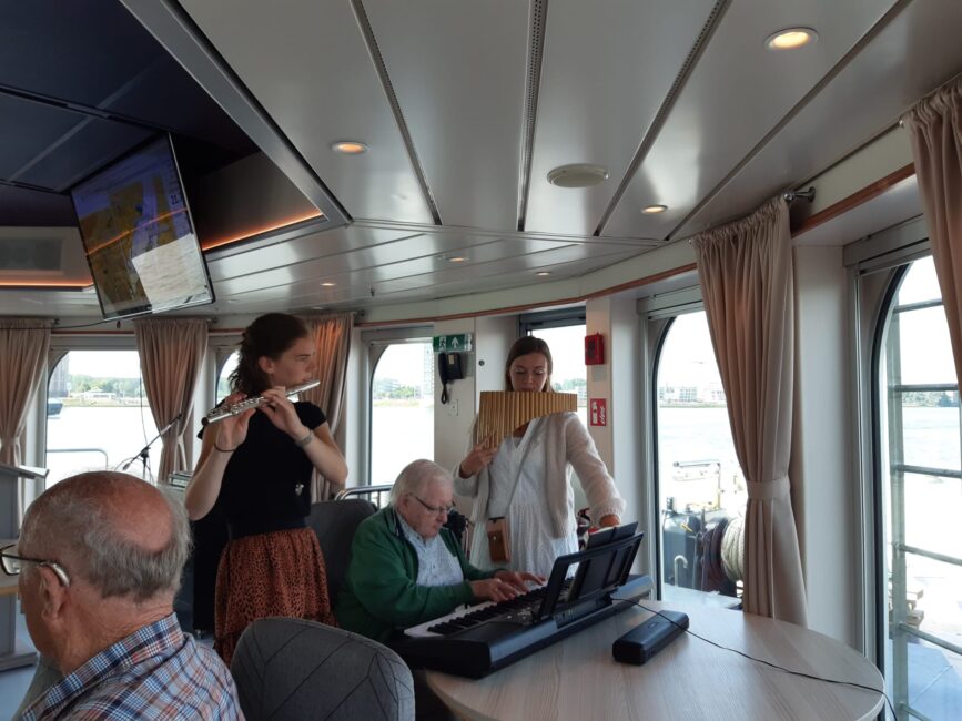 Muzikanten spelen fluit en keyboard voor een groep mensen in een boot met grote ramen en gordijnen, met uitzicht op het water.