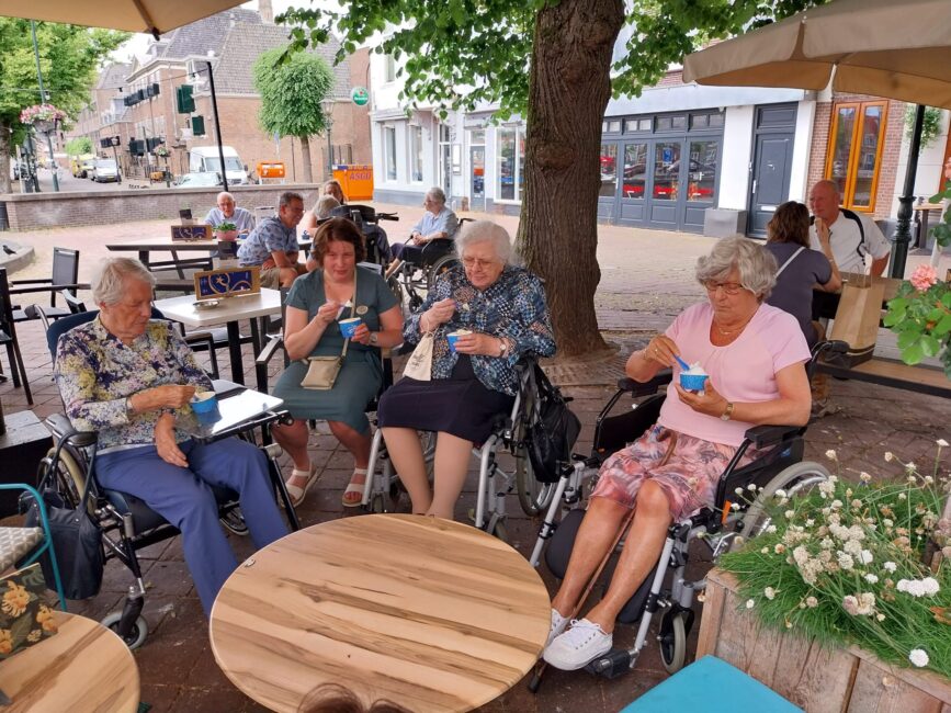 Vier oudere vrouwen zitten in rolstoelen onder een parasol op een terras en genieten van ijsjes op een zonnige dag.
