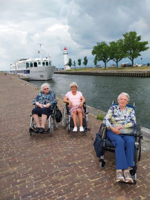Drie oudere vrouwen in rolstoelen zitten op een kade met een vuurtoren en een passerend schip op de achtergrond onder een bewolkte hemel.