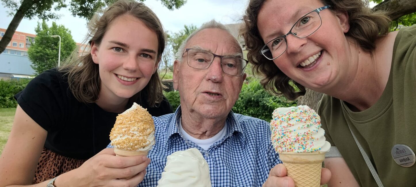 Drie mensen genieten van ijsjes in een park. De oudere man in het midden houdt een softijsje vast, terwijl de vrouwen hoorntjes hebben.