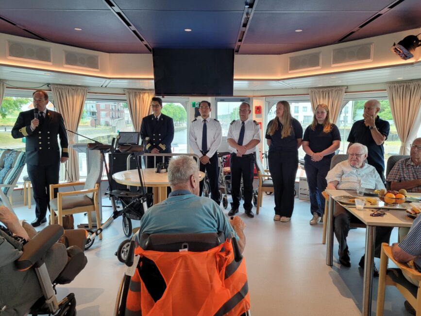 Groep mensen in uniformen en casual kleding staan voor een groep ouderen in een zaal met grote ramen en tafels.