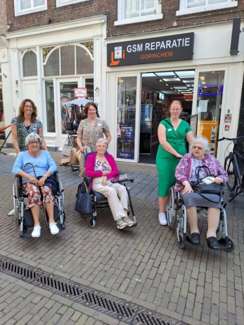 Groep van drie oudere vrouwen in rolstoelen en twee begeleiders poseren voor een winkel met een bord "GSM Reparatie".