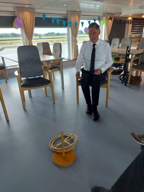 Man in een witte overhemd en zwarte stropdas zit in een kamer met stoelen en feestdecoraties, kijkend naar een houten object.