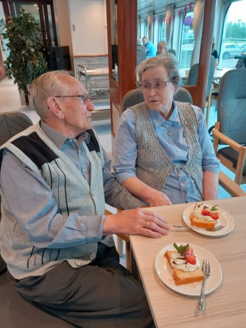 Een oudere man en vrouw zitten aan een tafel in een café, met voor hen borden met gebakjes, in een gezellige sfeer.