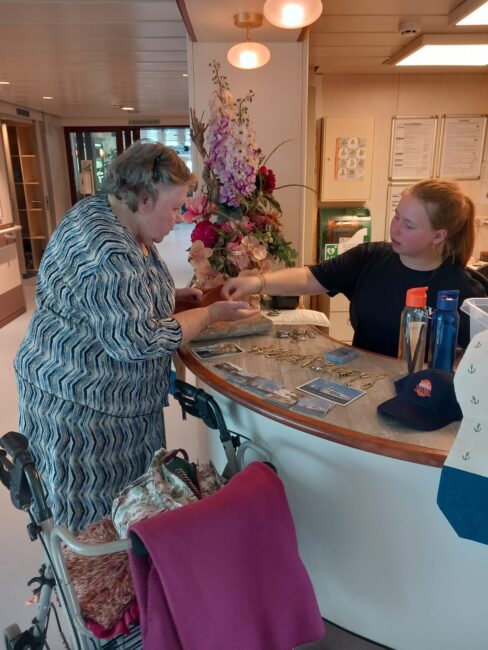 Een vrouw met een rollator praat met een medewerker achter een balie, met bloemen op de achtergrond in een verzorgingshuis.