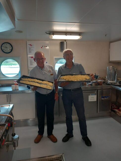 Twee mannen in een keuken houden elk een schaal met een gerecht vast. Er hangt een klok en een rond raam is zichtbaar.