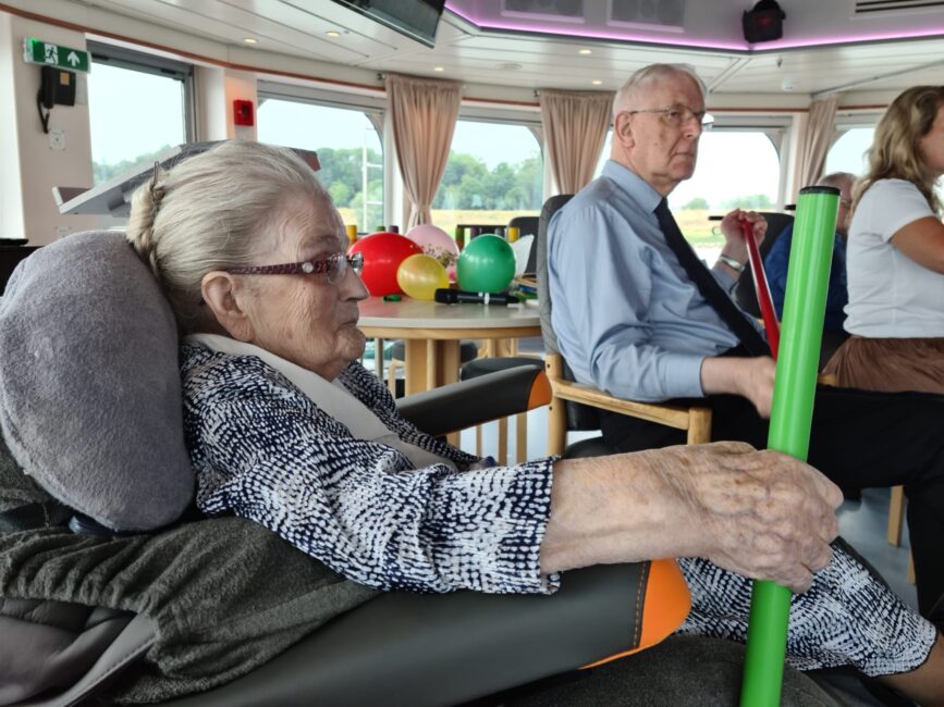 Oudere vrouw en man zitten in een comfortabele ruimte met grote ramen. De vrouw houdt een groene buis vast.