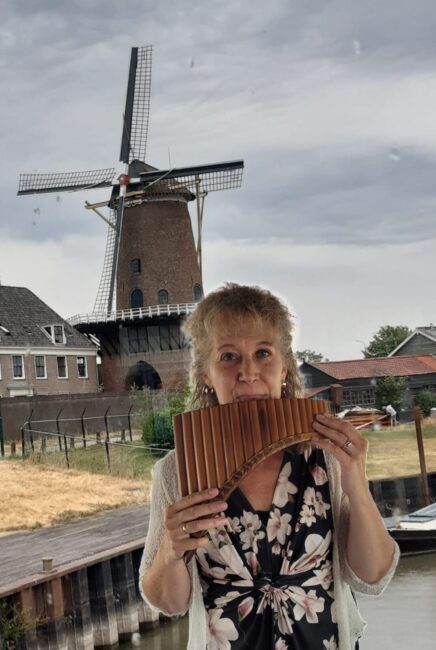 Vrouw speelt panfluit voor een traditionele Nederlandse windmolen, met huizen en een kanaal op de achtergrond.