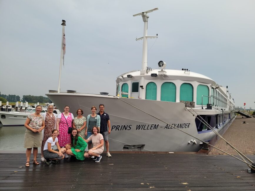 Groep mensen poseert voor een groot wit schip genaamd "Prins Willem-Alexander" aan een rustige kade op een bewolkte dag.