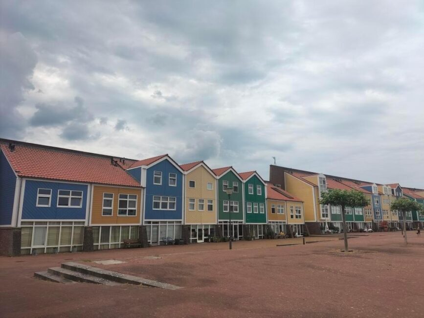 Rij van kleurrijke huizen met puntdaken onder een bewolkte hemel. De gevels zijn blauw, geel en groen. Lege straat op de voorgrond.