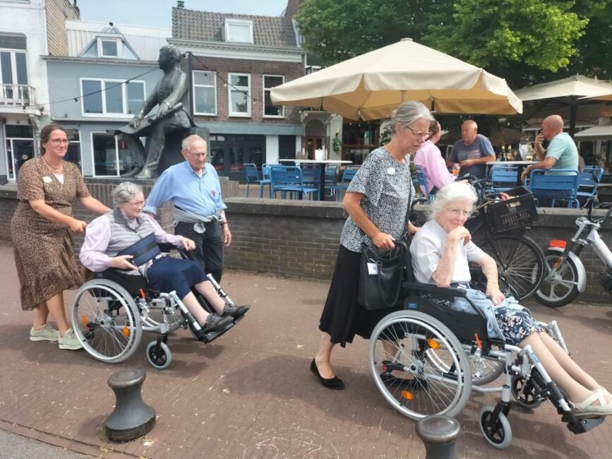 Groep mensen duwt ouderen in rolstoelen over een trottoir. Op de achtergrond zijn een terras en een standbeeld zichtbaar.
