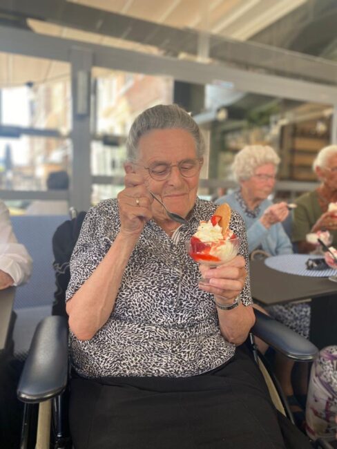 Een oudere vrouw geniet van een ijsje in een coupe, zittend aan een tafel in een café, omringd door andere mensen.