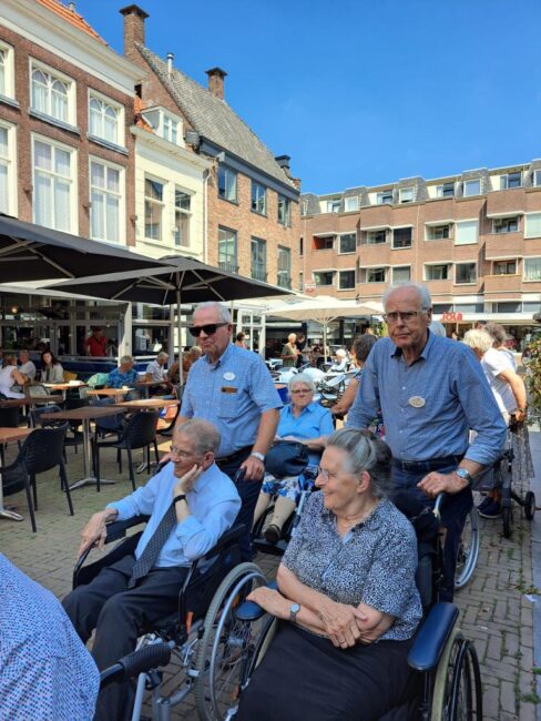 Een groep mensen in rolstoelen wordt begeleid door verzorgers op een zonnig plein met terrassen en historische gebouwen.