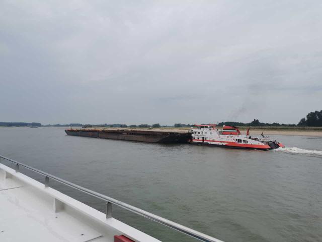Een rode en witte sleepboot trekt een groot, lang schip over een brede rivier onder een bewolkte hemel. Land is zichtbaar in de verte.