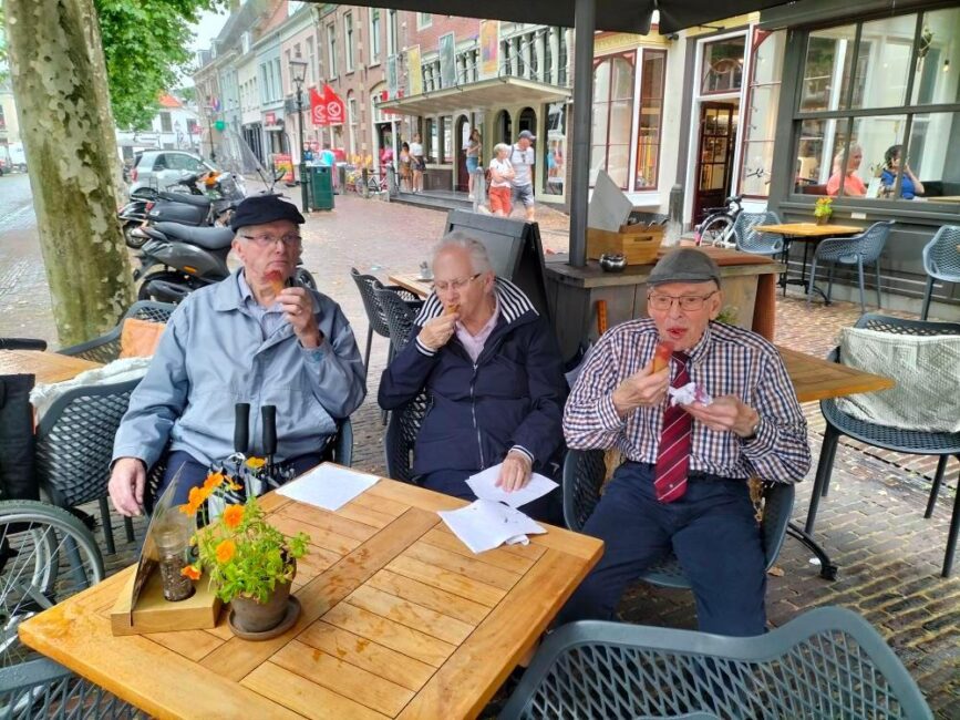 Drie oudere mannen zitten buiten aan een tafel bij een café en eten ijsjes. Op de achtergrond zijn winkels en fietsen te zien.