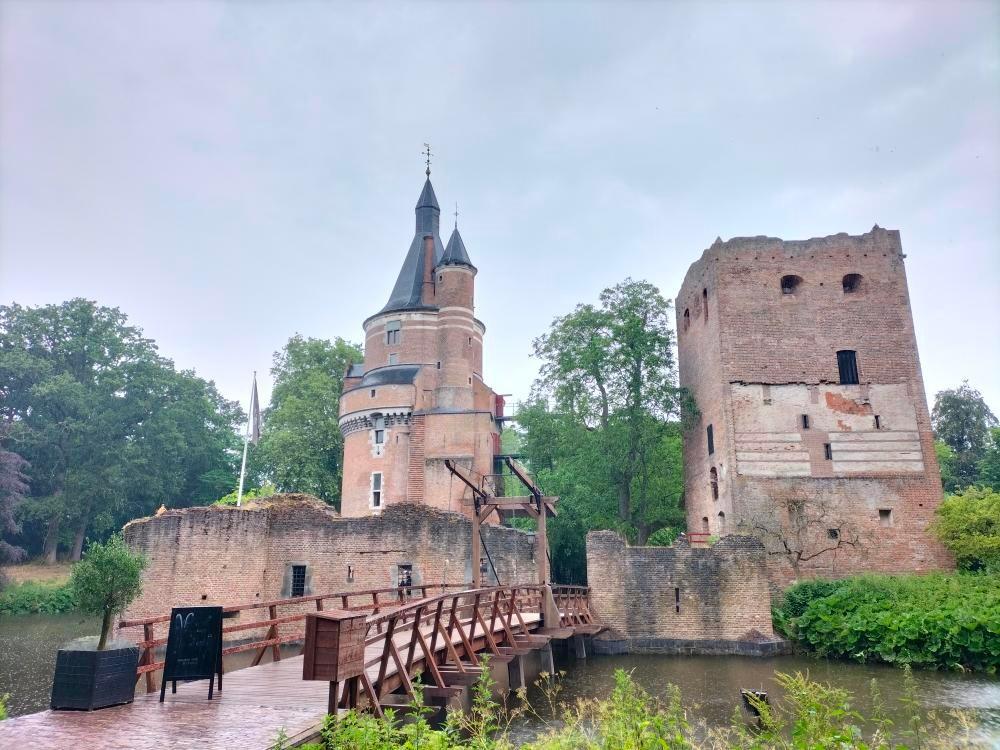 Middeleeuws kasteel met toren en ruïne, omgeven door groen en water. Een houten brug leidt naar de ingang van het kasteel.