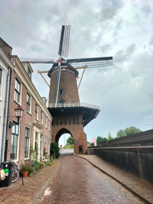 Een historische windmolen naast een smalle, geplaveide straat met bakstenen gebouwen en een bewolkte lucht op de achtergrond.