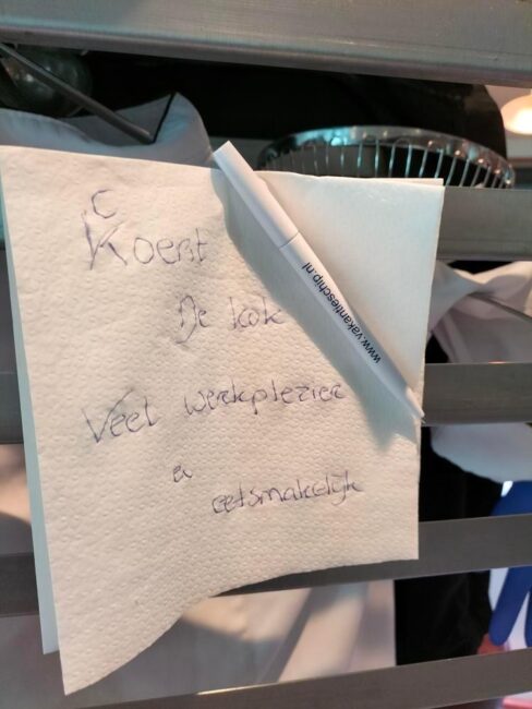 Een servet met handgeschreven tekst hangt aan een rek, met een pen erboven. De tekst is deels leesbaar en informeel.