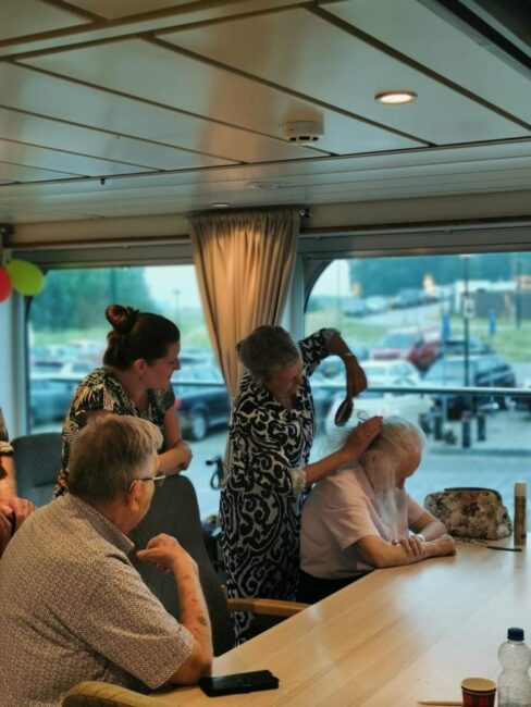 Een groep mensen zit aan een tafel in een kamer met grote ramen. Een vrouw staat en lijkt een andere vrouw te kappen.