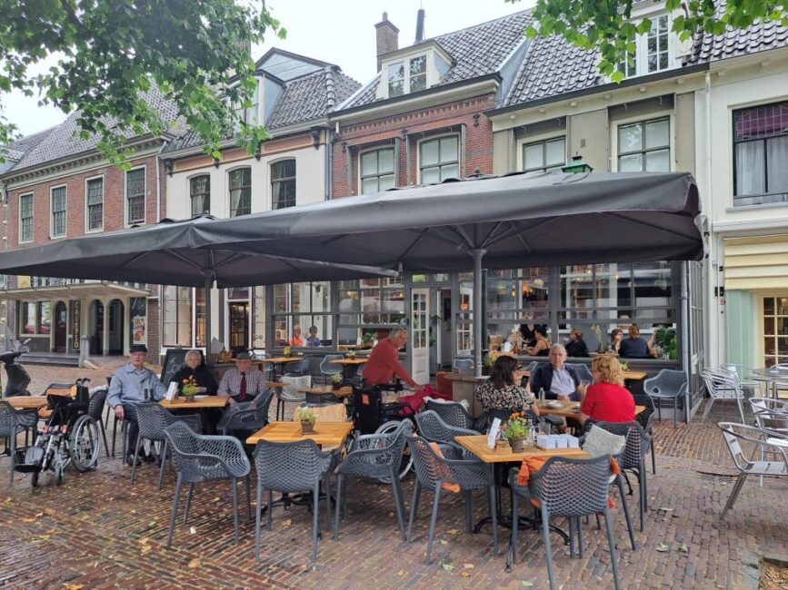 Terras met mensen die aan tafels zitten onder een grote parasol, omringd door historische gebouwen en groene bomen.
