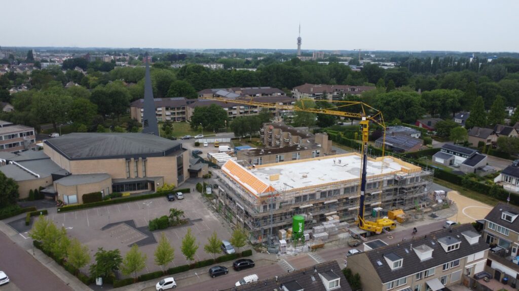 Luchtfoto van een bouwplaats in een woonwijk met een hijskraan en een gebouw in aanbouw, omgeven door bomen en huizen.