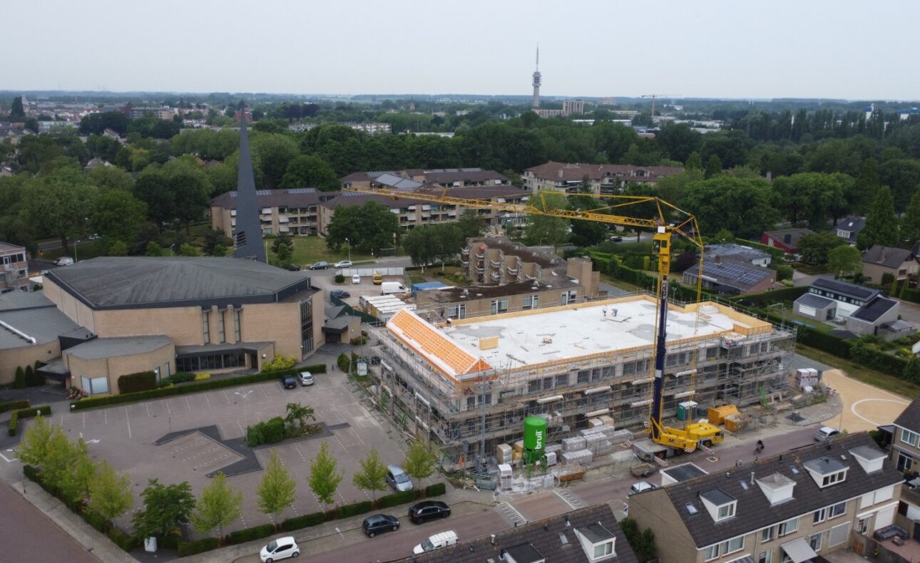 Luchtfoto van een bouwplaats in een woonwijk met een hijskraan en een gebouw in aanbouw, omgeven door bomen en huizen.