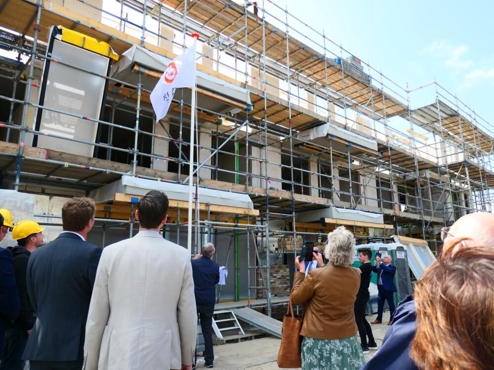 Groep mensen kijkt naar een gebouw in aanbouw met steigers en een vlag. Enkele personen dragen bouwhelmen.