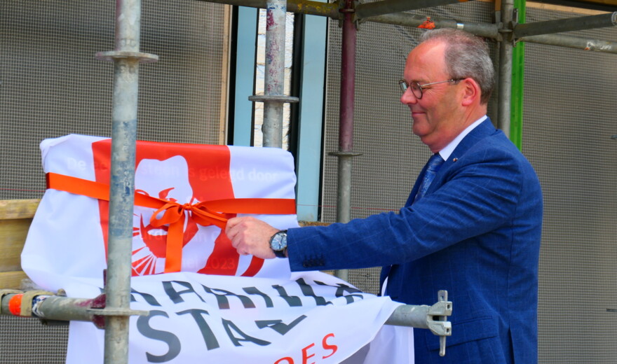 Man in blauw pak onthult een bord met een rood-wit logo en tekst, omringd door steigerbuizen.