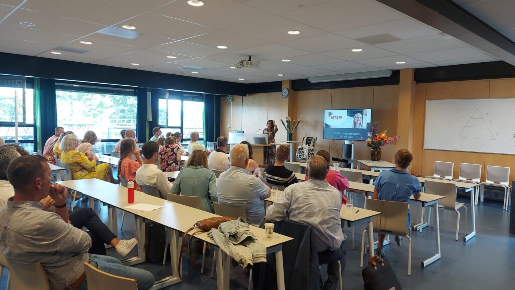 Groep mensen zit in een vergaderruimte en kijkt naar een presentatie op een scherm. De ruimte heeft grote ramen en een whiteboard.
