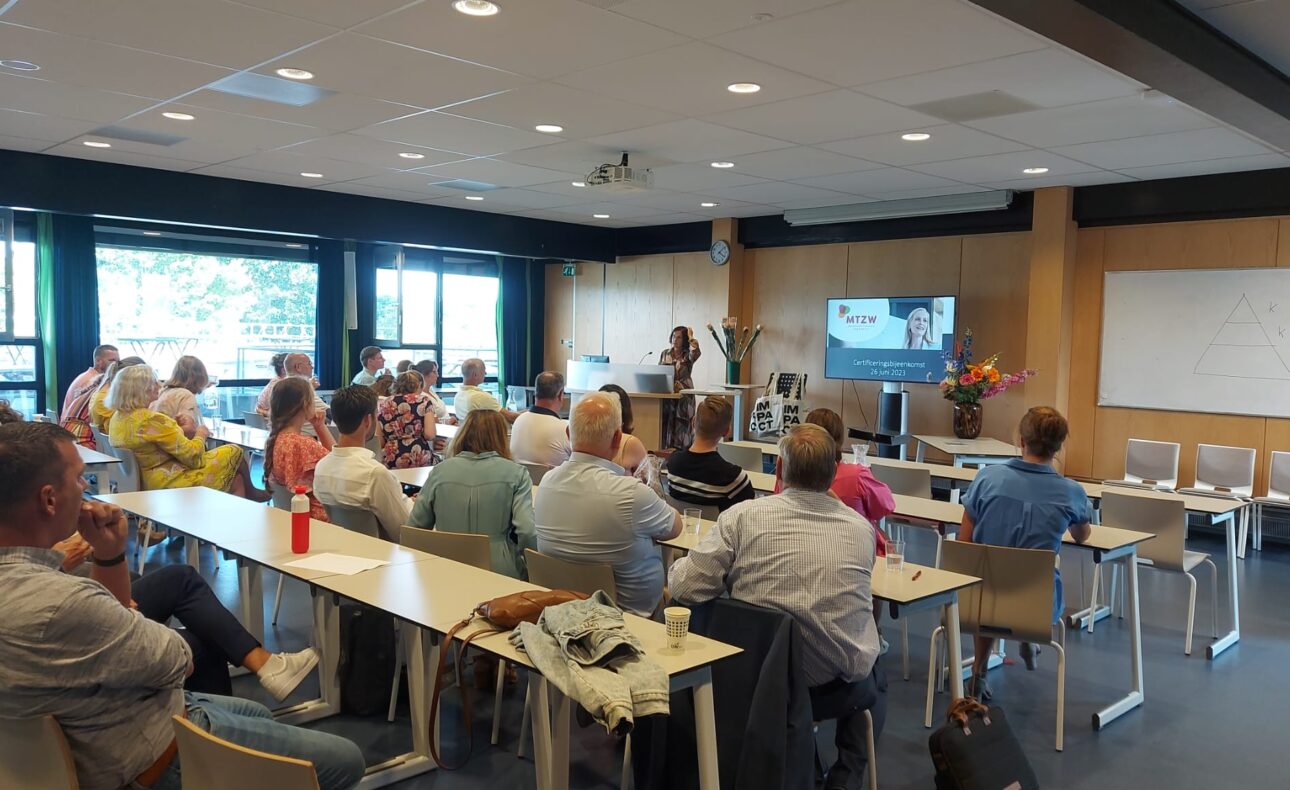 Groep mensen zit in een vergaderruimte en kijkt naar een presentatie op een scherm. De ruimte heeft grote ramen en een whiteboard.