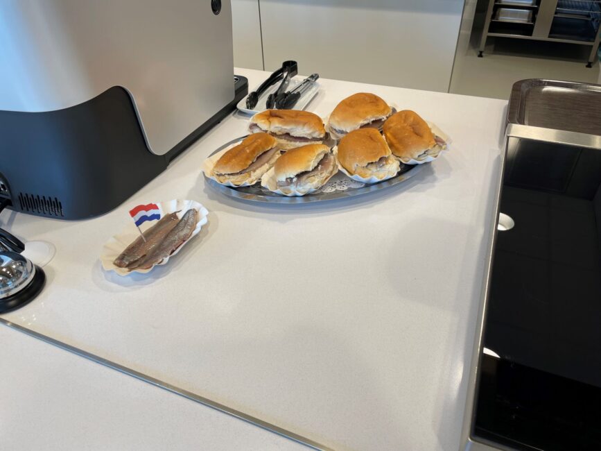 Bord met broodjes haring en een schaaltje met haring en Nederlandse vlaggetjes op een witte tafel in een keuken.