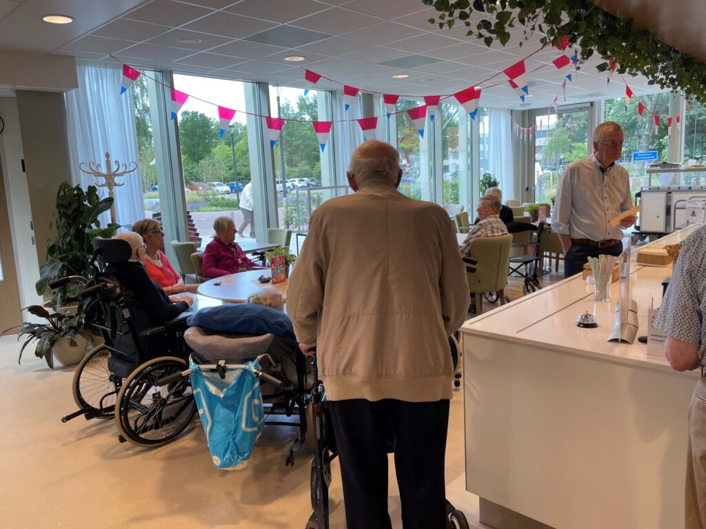 Een groep ouderen geniet van een haringmiddag in een lichte ruimte met versieringen. Mensen zitten aan tafels en praten.