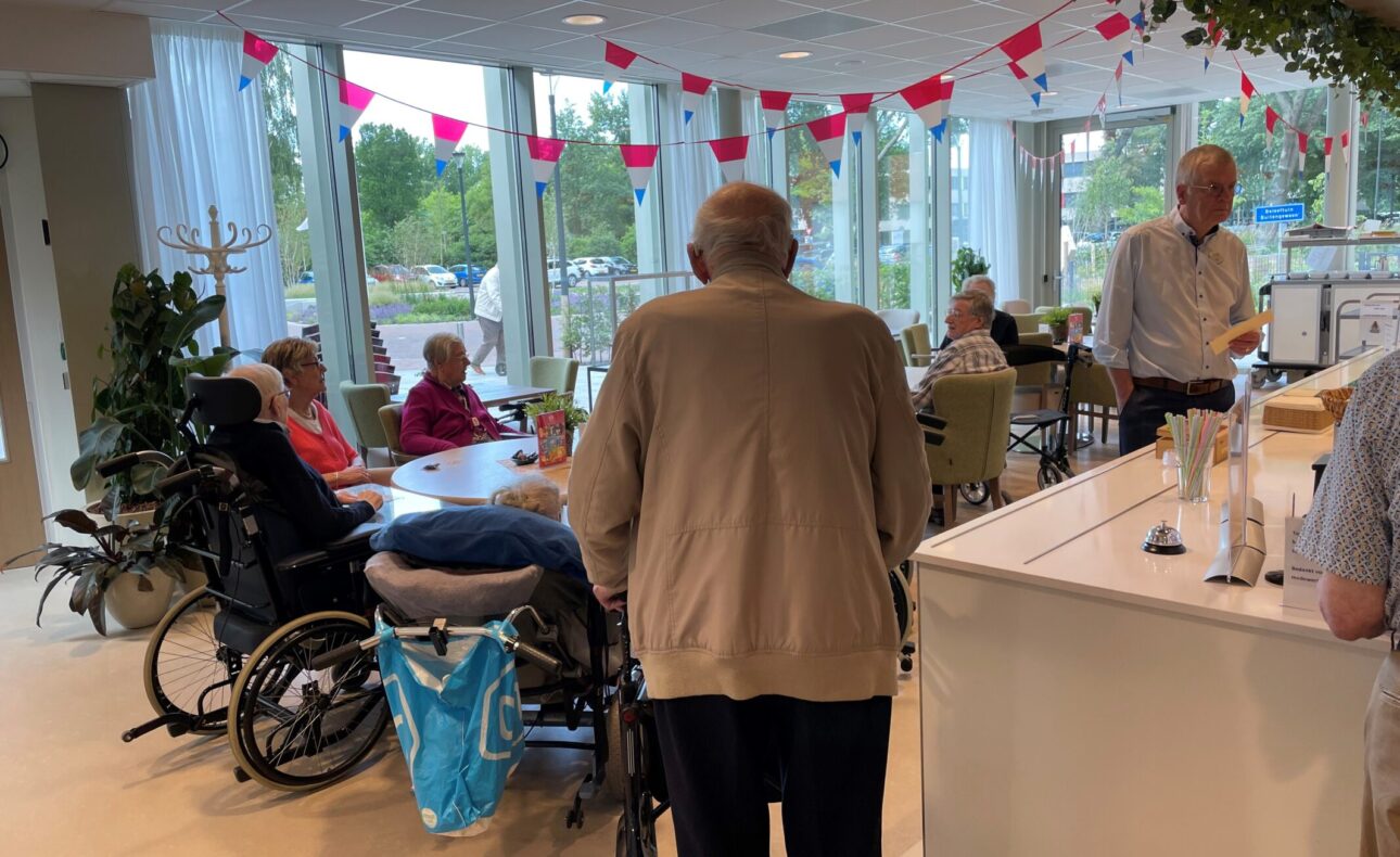 Een groep ouderen geniet van een haringmiddag in een lichte ruimte met versieringen. Mensen zitten aan tafels en praten.