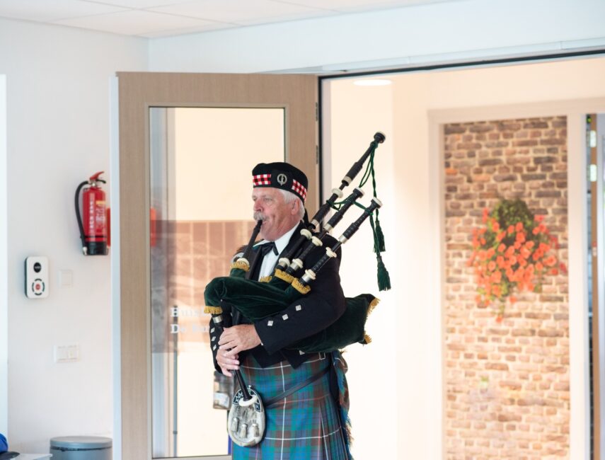 Een doedelzakspeler in traditionele Schotse kleding speelt binnen, met een open deur en een bakstenen muur op de achtergrond.