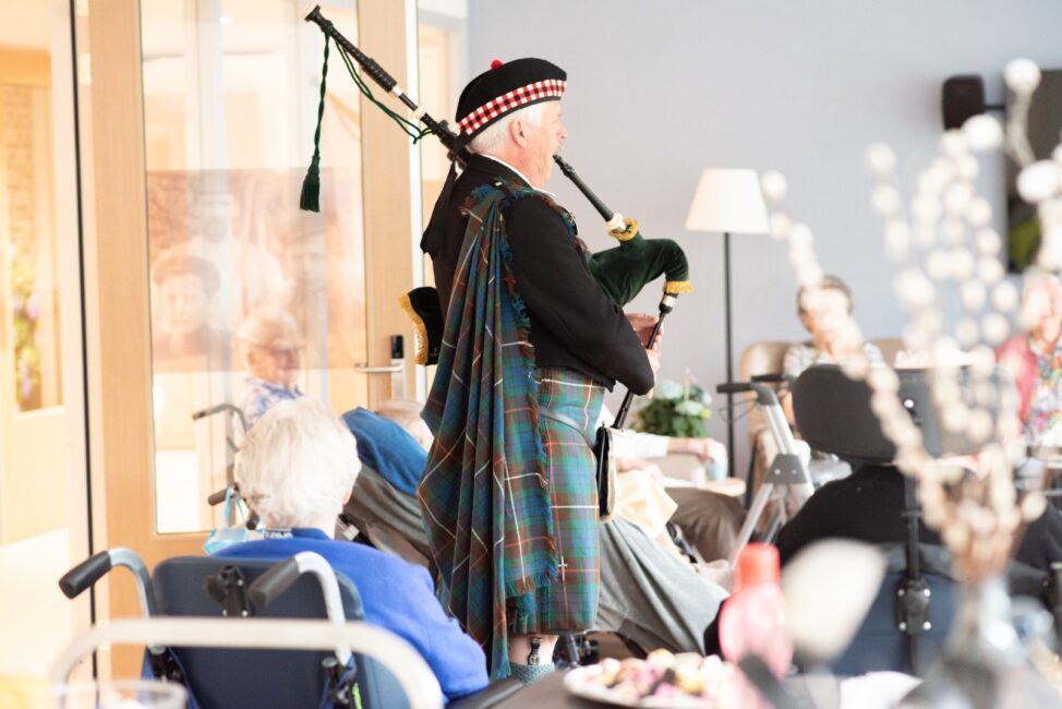 Een man in traditionele Schotse kleding speelt doedelzak voor een groep mensen in een lichte kamer met rolstoelen.