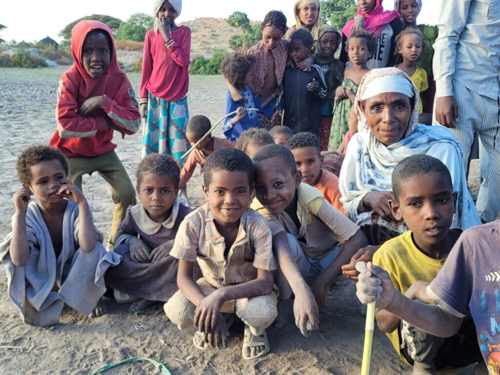 Groep kinderen en een volwassene poseren buiten op een zandweg. Ze lachen en kijken naar de camera, met heuvels op de achtergrond.