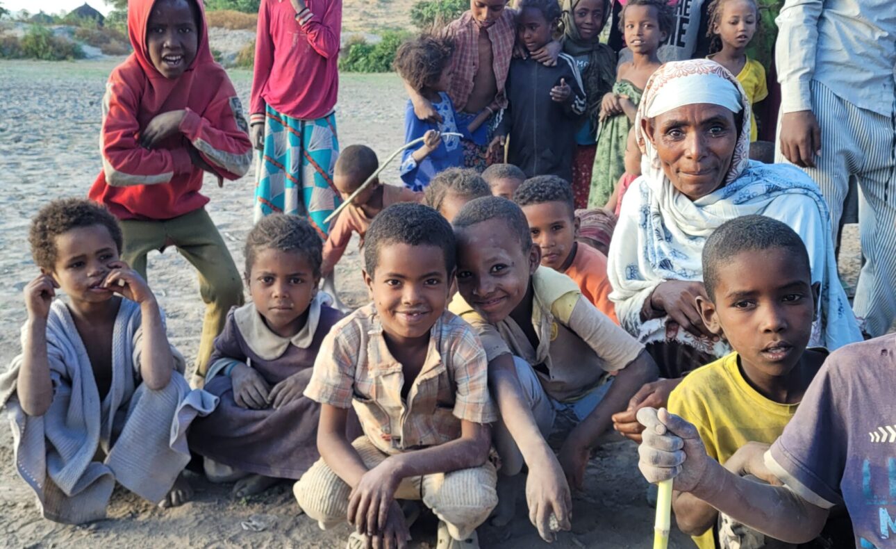 Groep kinderen en een volwassene poseren buiten op een zandweg. Ze lachen en kijken naar de camera, met heuvels op de achtergrond.