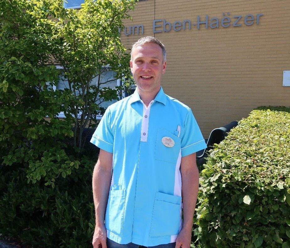 Man in lichtblauw uniform staat buiten voor een gebouw met de naam "Eben-Haëzer". Groene struiken en bakstenen muur op achtergrond.