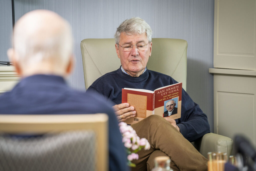 Een oudere man leest een boek terwijl hij in een comfortabele stoel zit. Op de achtergrond is een vaas met bloemen zichtbaar.