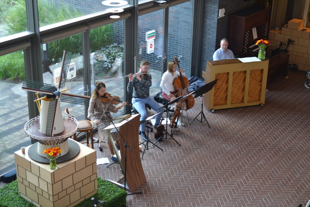 Muzikanten spelen viool, fluit, cello en piano in een lichte ruimte met grote ramen en een decoratieve molen op de voorgrond.