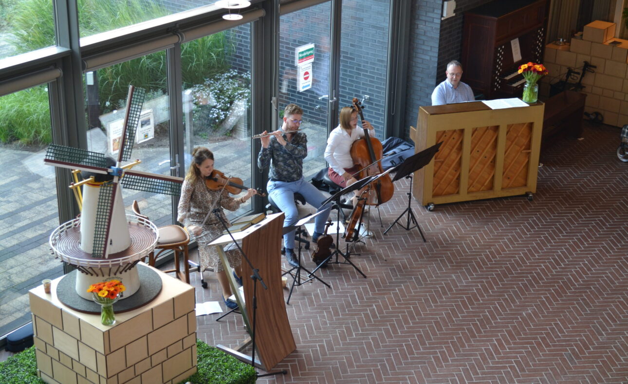 Muzikanten spelen viool, fluit, cello en piano in een lichte ruimte met grote ramen en een decoratieve molen op de voorgrond.